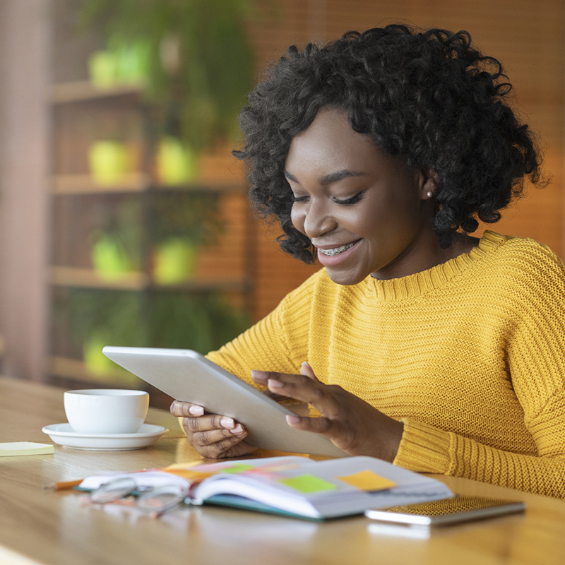 Woman using a touchscreen tablet, smiling with coffee and notebook.