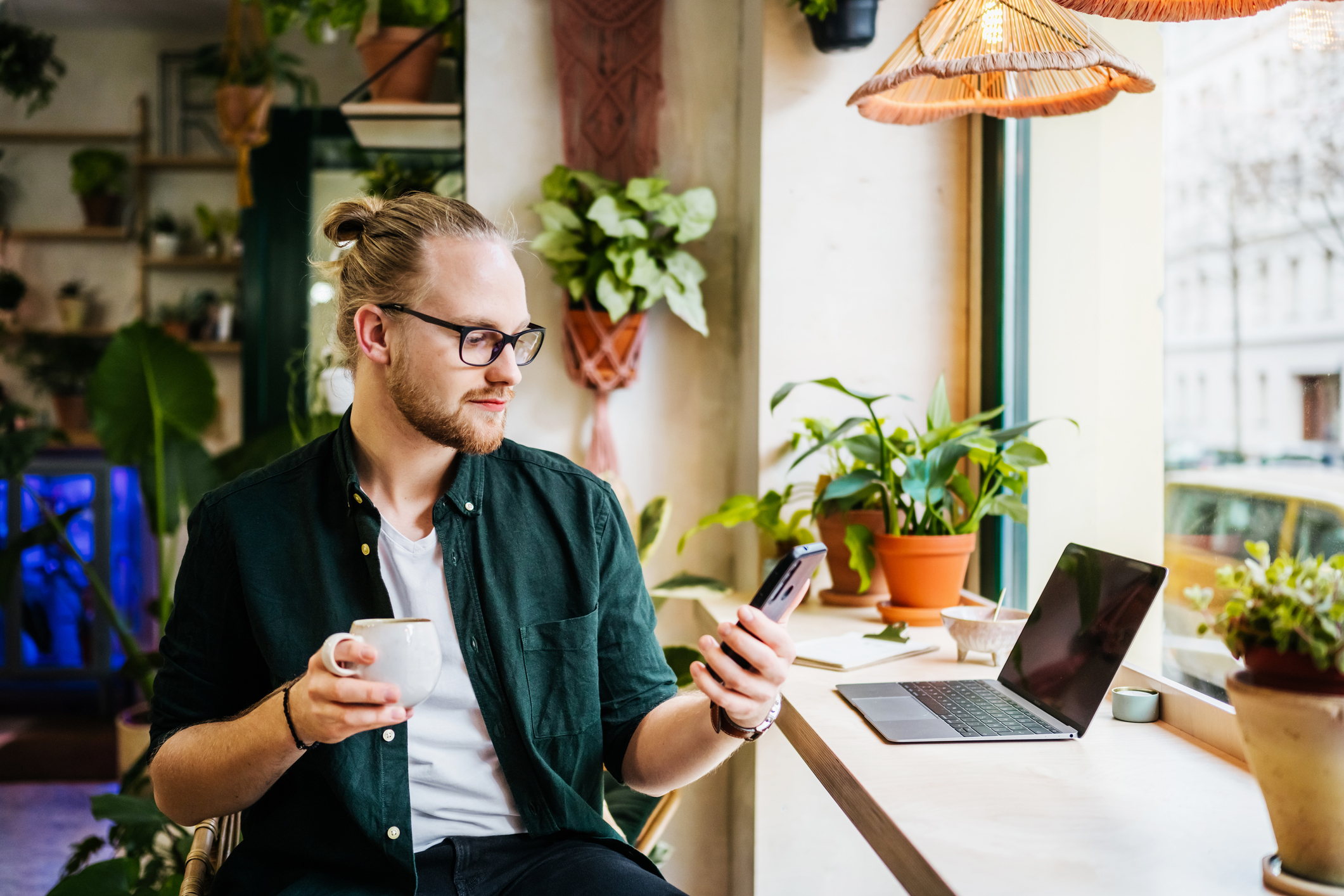 Homem sentado em uma mesa de escritório em casa, em frente a um laptop, segurando uma xícara de café.