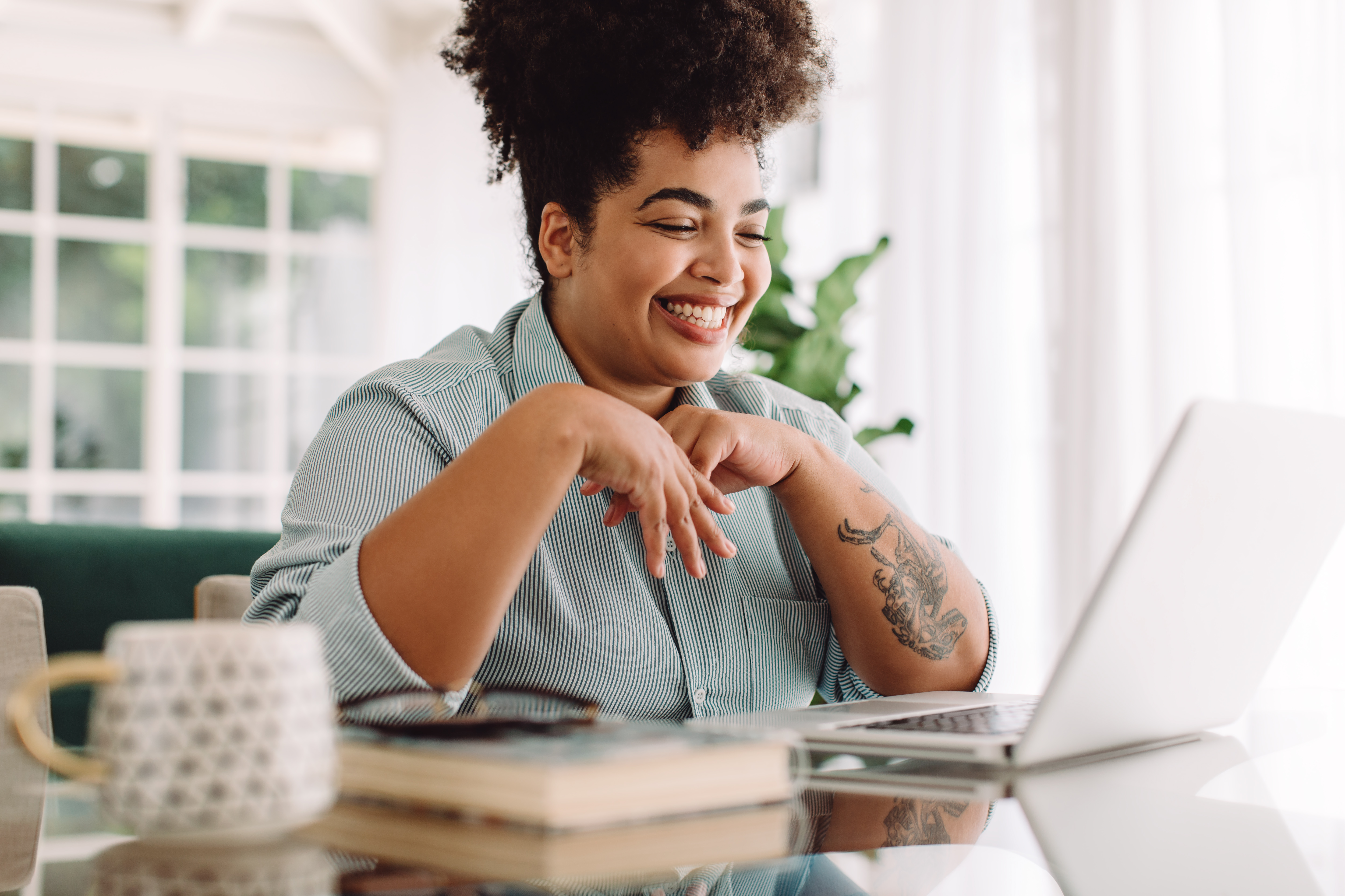 Uma pessoa sorridente está usando um laptop, uma caneca e um livro estão por perto.