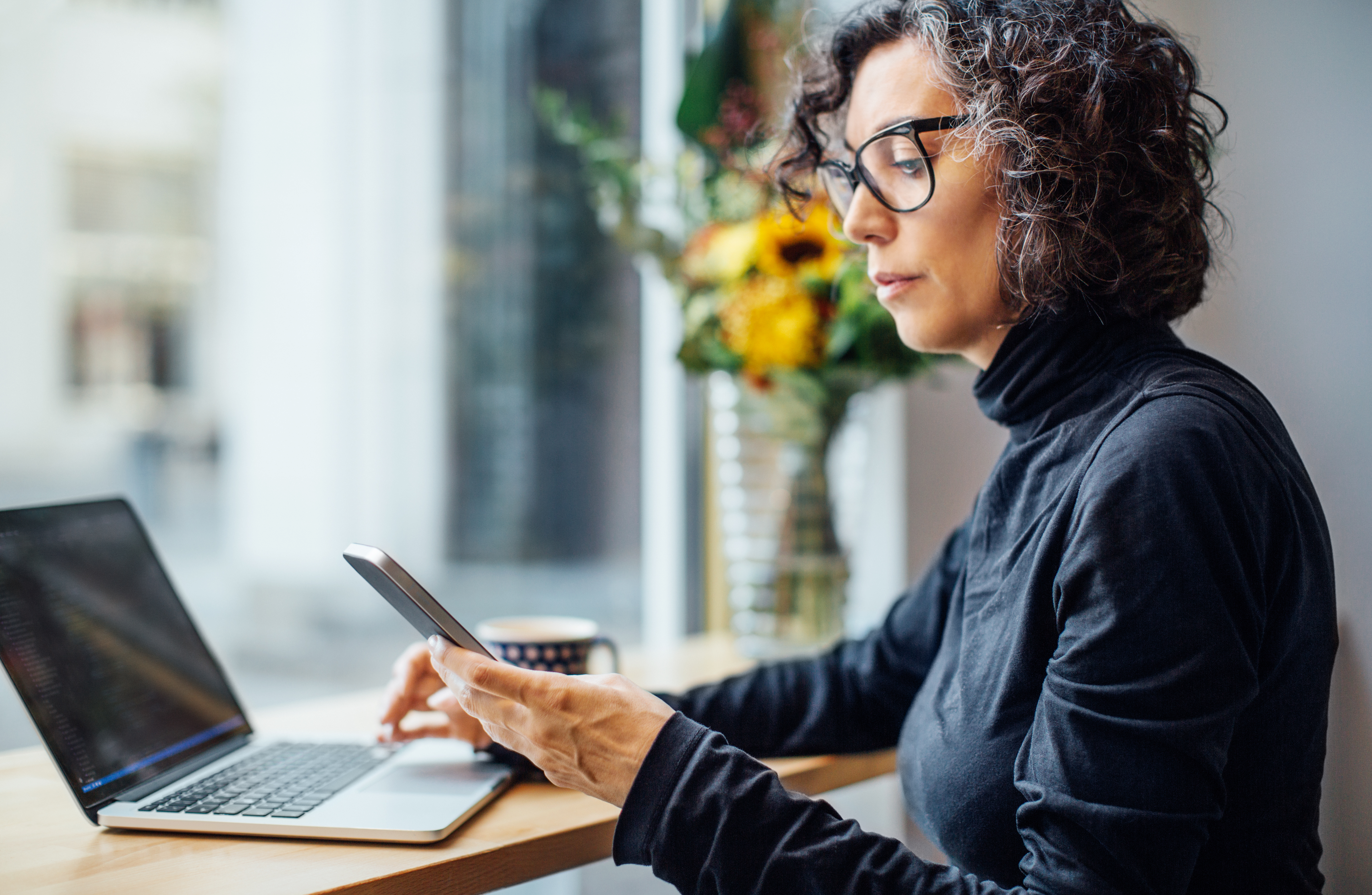 Mulher usando um smartphone e um laptop em um café; online e presencial.