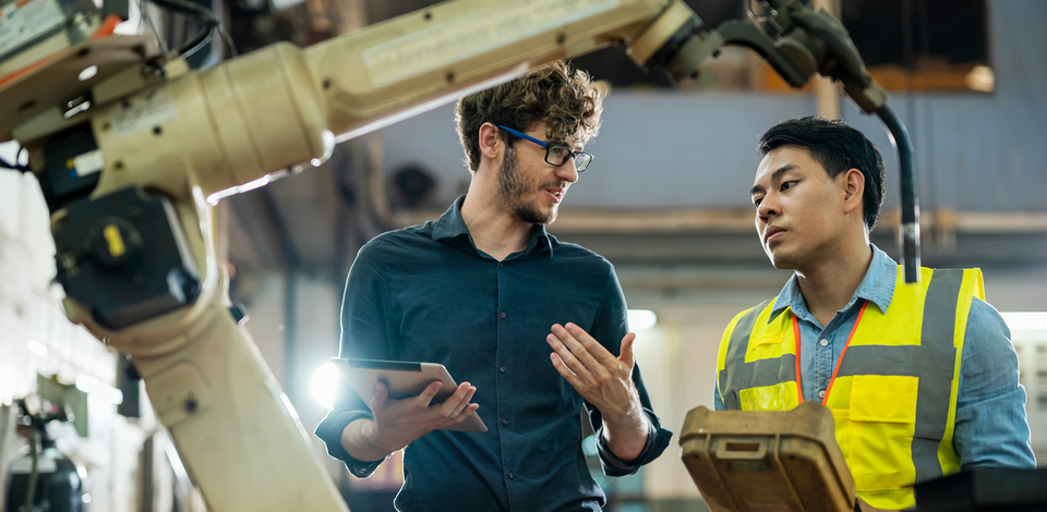 Two engineers, one holding a tablet, discuss a robotic arm.