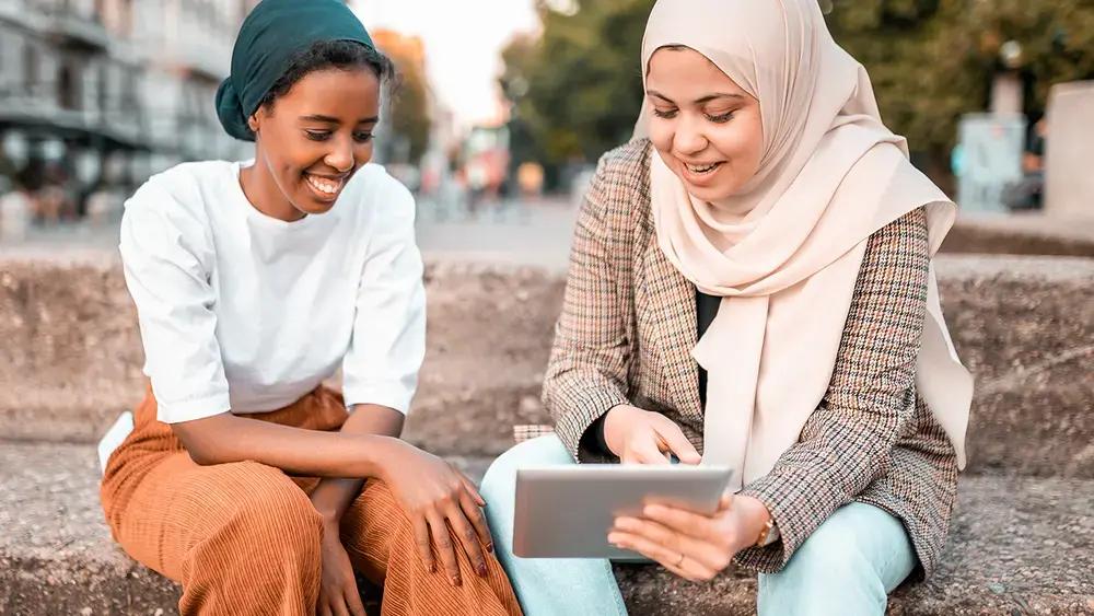 Two smiling women sit outdoors, looking at a tablet.
