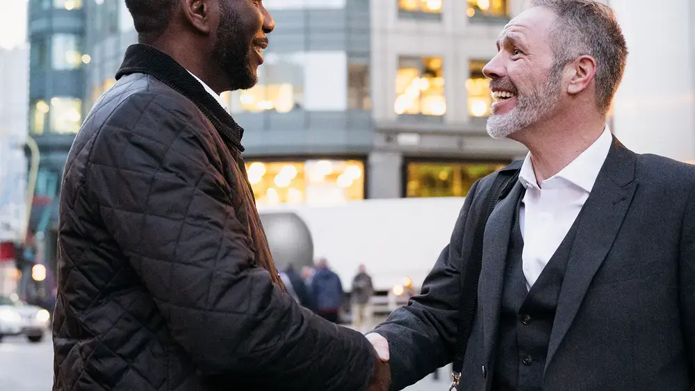 Two smiling coworkers shaking hands outdoors.
