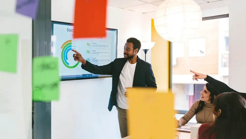 A man leads a team meeting, presenting data on a screen.
