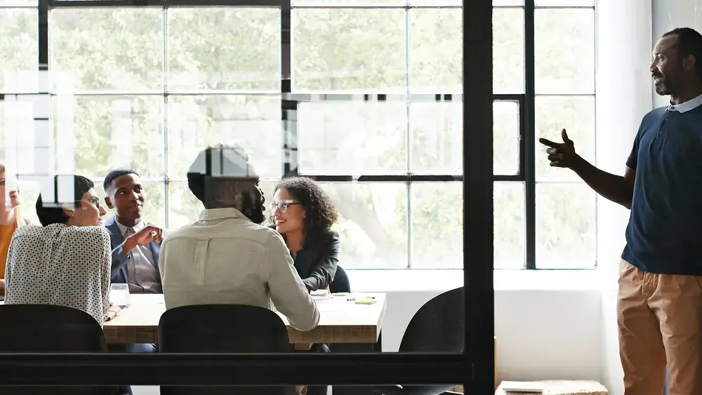 A presenter speaks to a group in a glass-walled conference room.