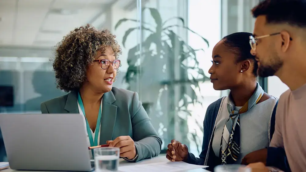 A woman discusses something with two people while looking at a laptop.