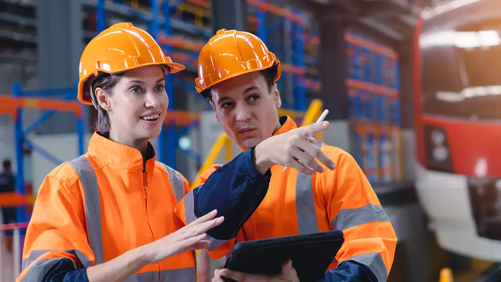 Two workers in orange vests review a tablet.