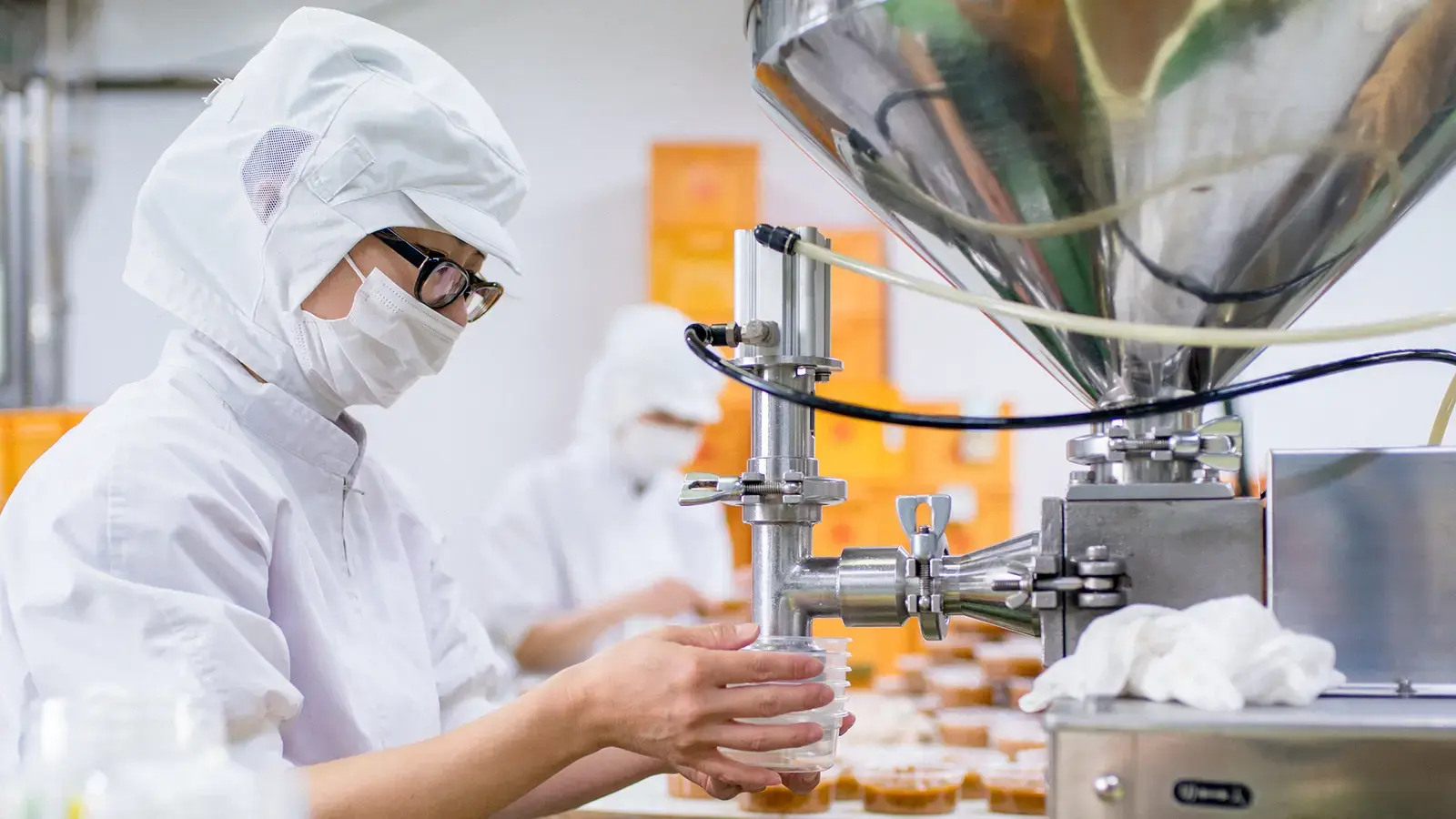 Workers with white caps, masks, lab coats work in a clean food preparation facility