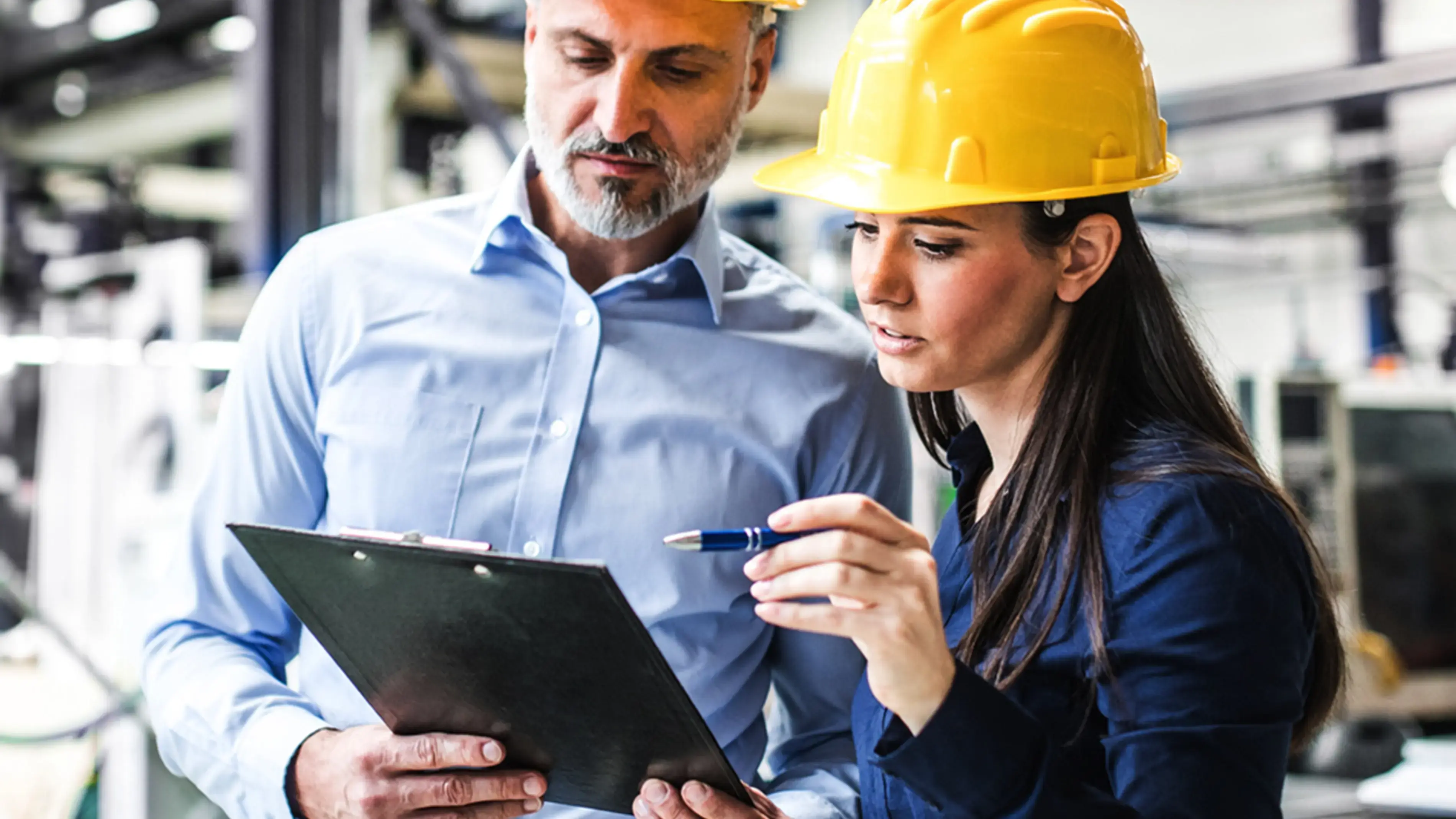 Two coworkers review a document on site, wearing yellow hard hats.