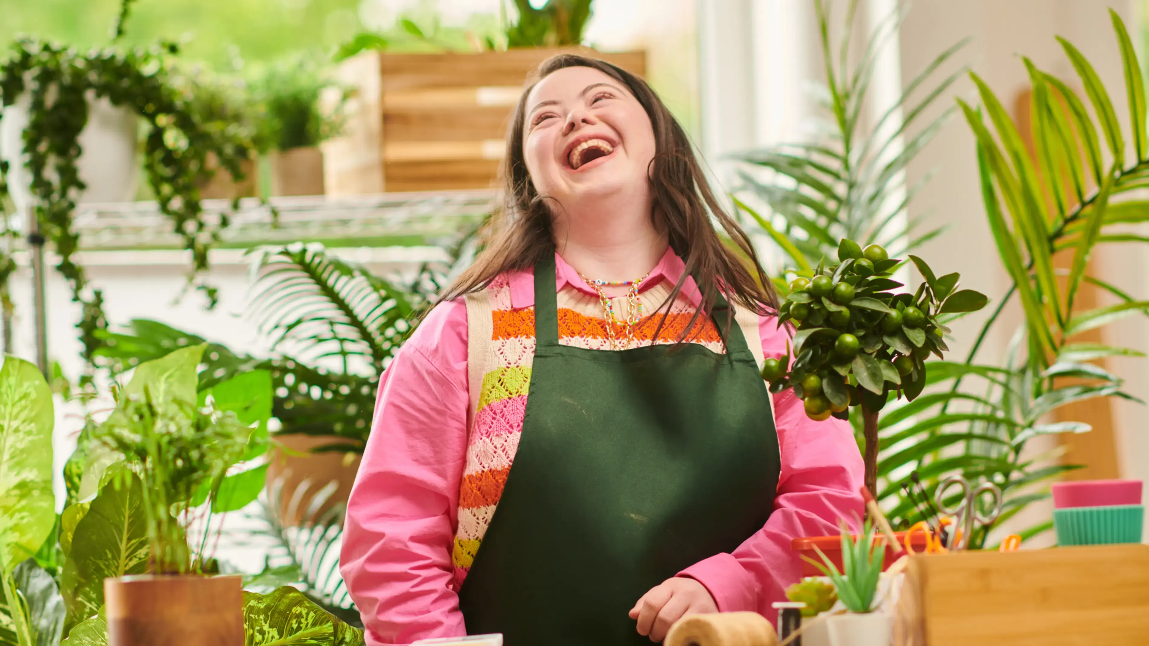 Uma mulher sorridente de camisa rosa, rodeada por plantas em uma empresa de médio porte.