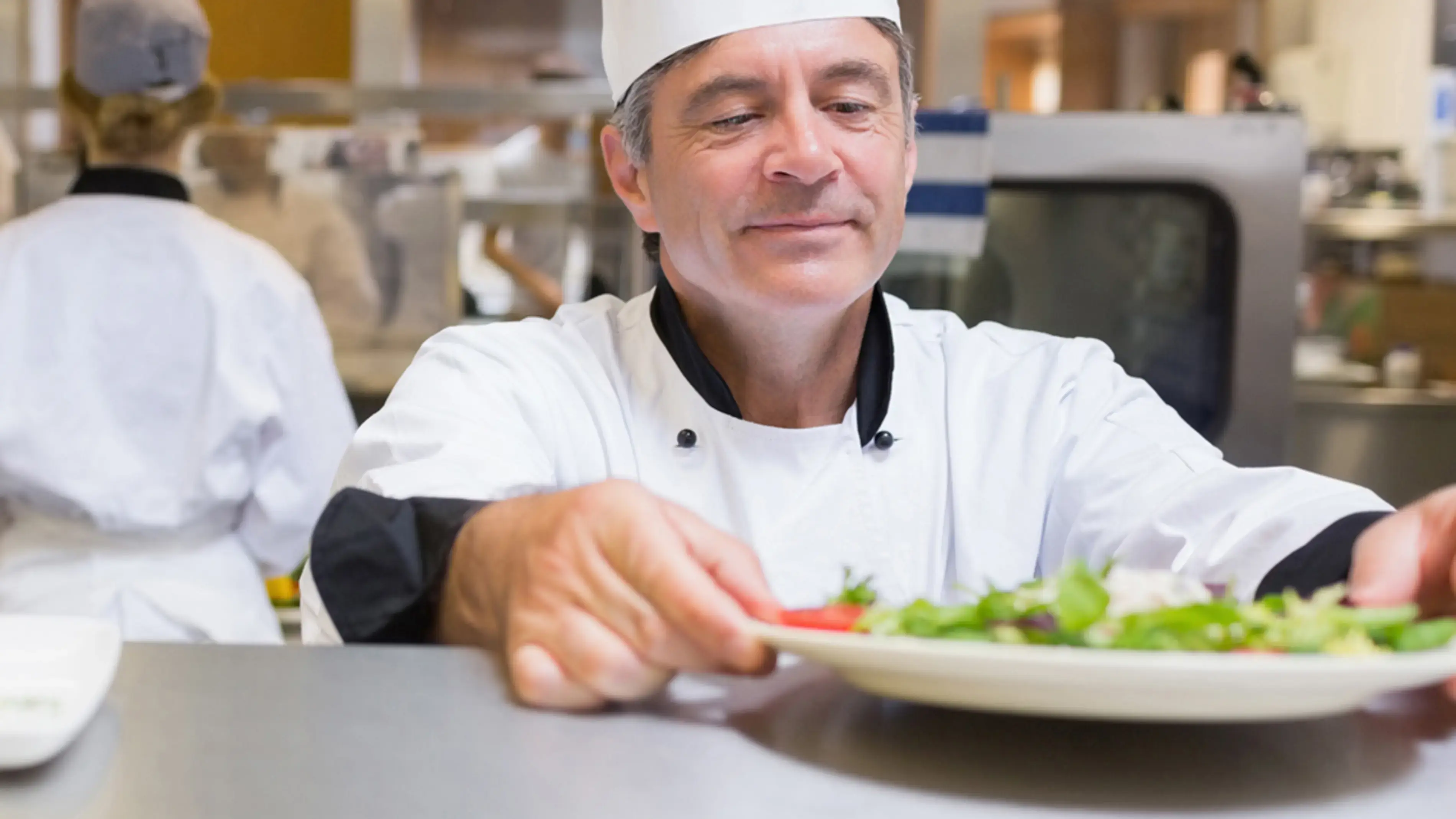 Um chef colocando um prato de comida, um colega de trabalho está ao fundo.