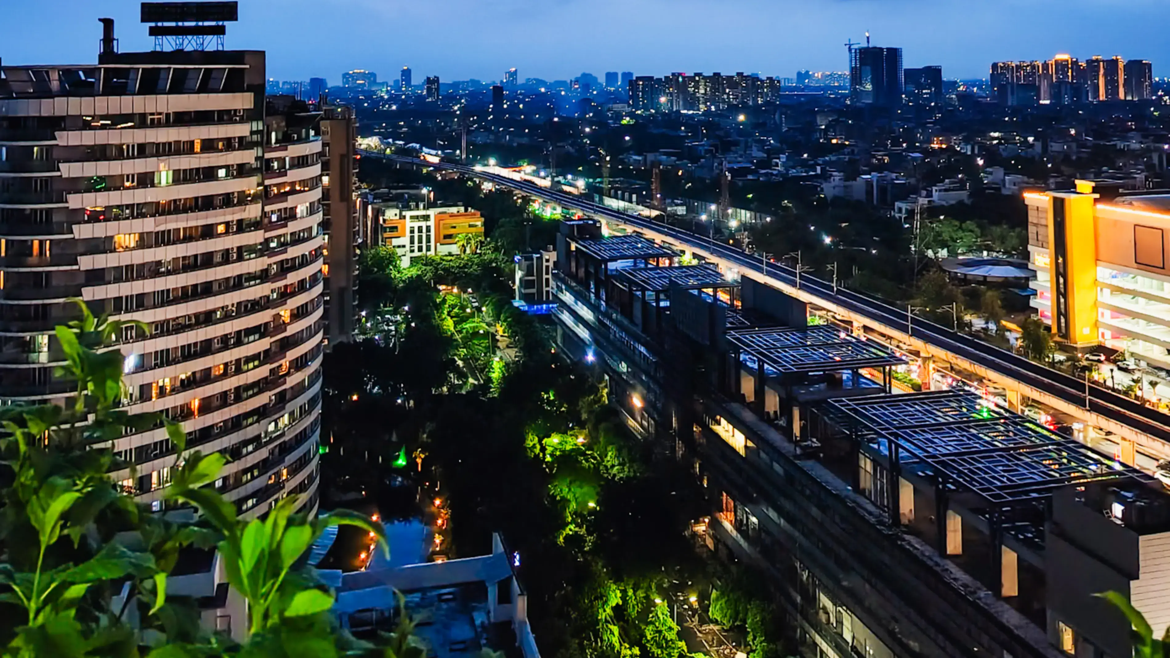 Photograph of a city skyline of Noida with buildings and a train track at dusk.