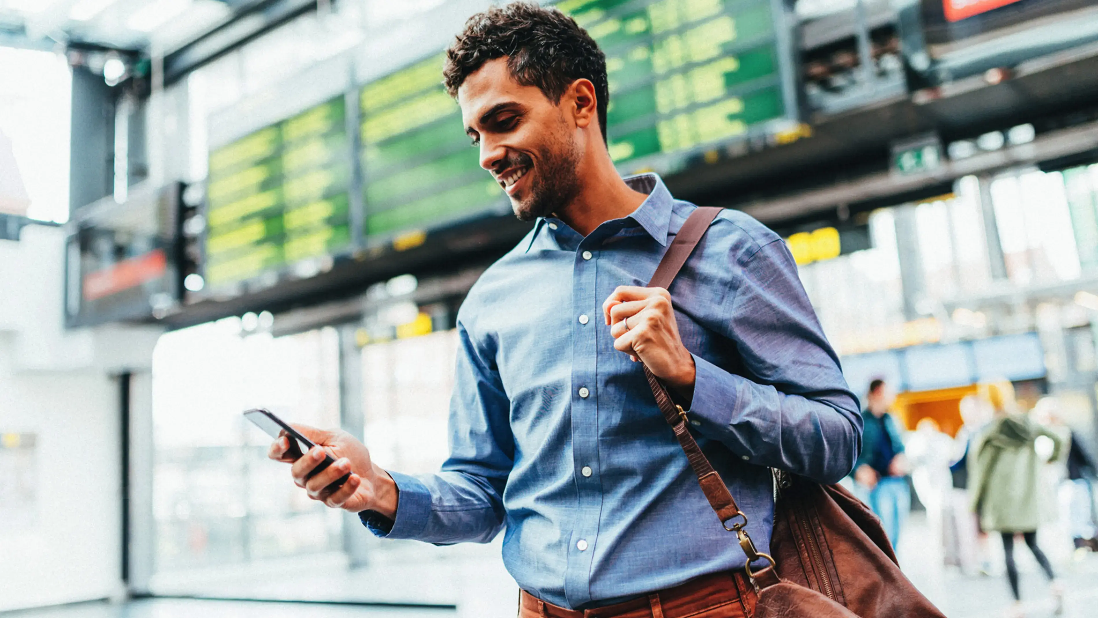 A smiling man using a smartphone, with a bag on his shoulder.