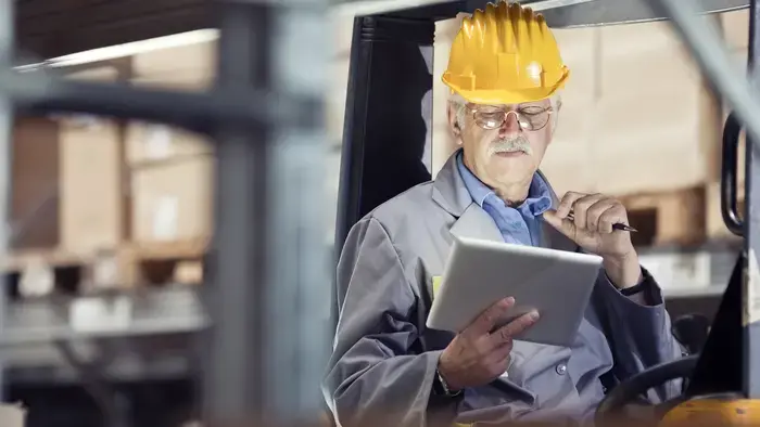 Older man in hardhat using tablet in forklift.
