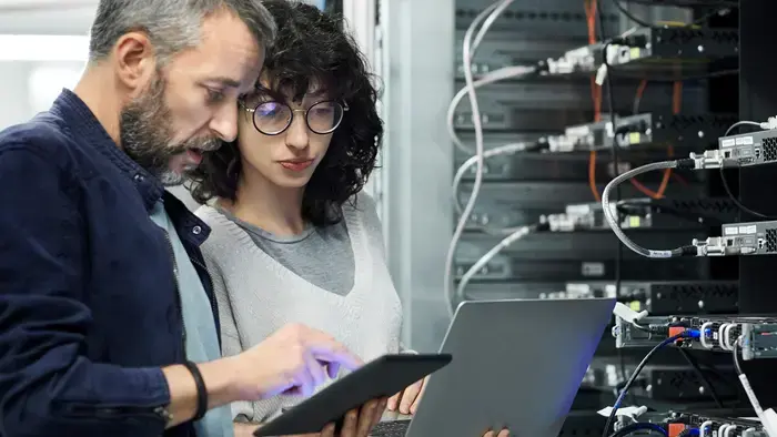 Two coworkers review data on a laptop and tablet near a server rack.
