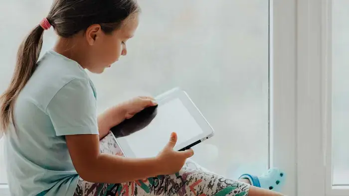 A child with a touchscreen device sitting by a window wearing green Crocs..