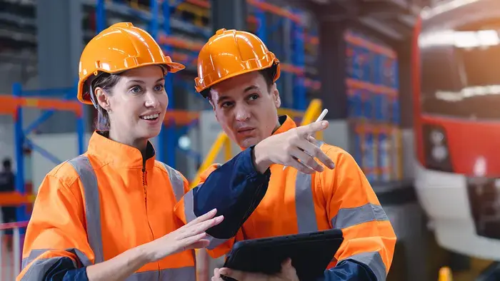 Two workers in orange vests review a tablet.