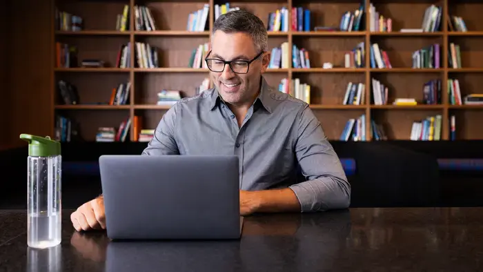 Homem de óculos sorri enquanto usa um laptop; fundo de estante.