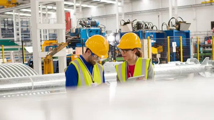 Duas pessoas com capacetes e coletes amarelos analisam dados em uma fábrica.