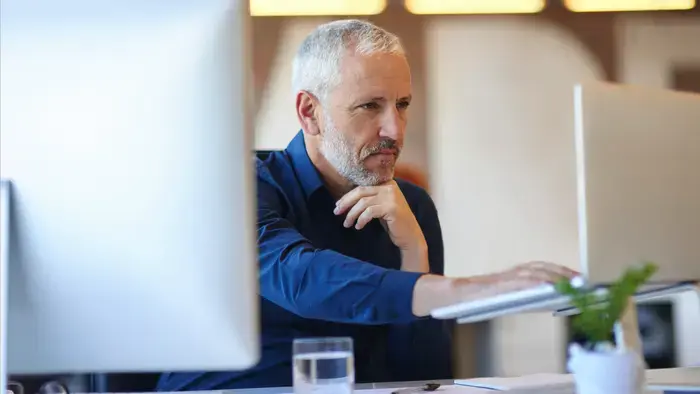 Um homem trabalha em um computador com uma planta pequena ao lado.