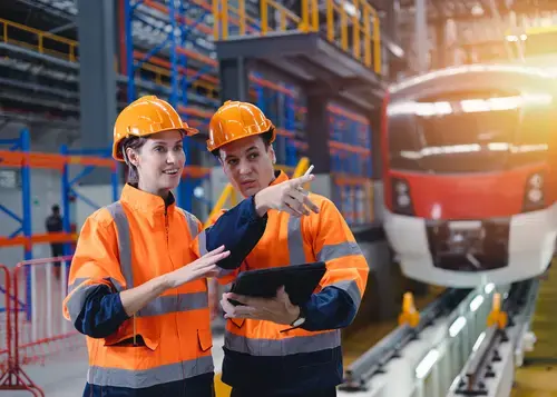 Two workers in orange vests review a tablet near a train.