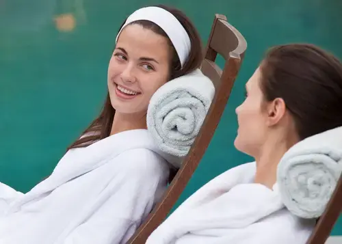 Women in white terry robes chat in lounge chairs while drinking cucumber water