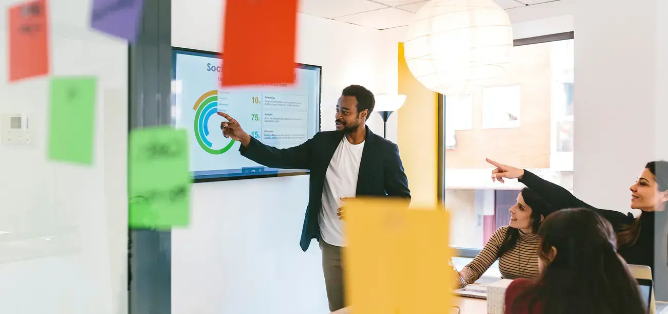 A man leads a team meeting, presenting data on a screen.
