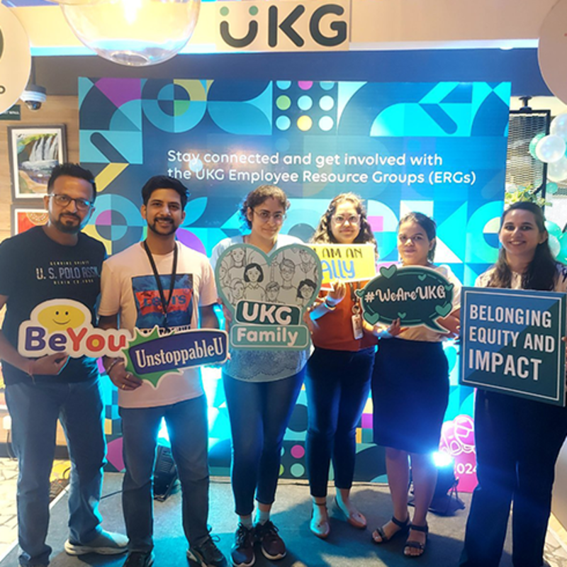 A group of six UKG employees are standing in front of a colorful backdrop