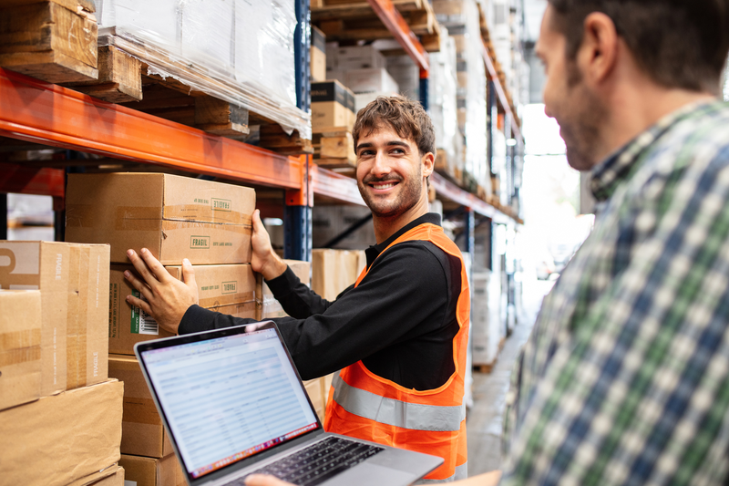 Two warehouse workers check inventory on a laptop while smiling at one another.