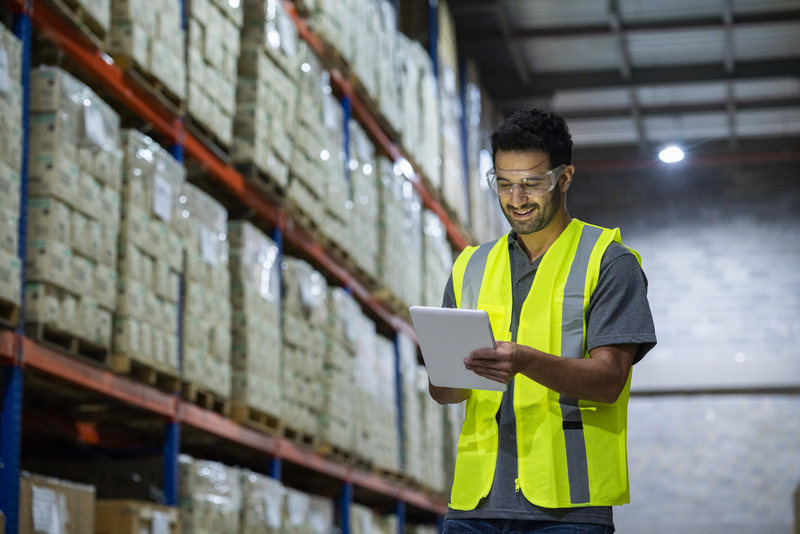 A man wearing a safety vest and glasses is looking at a tablet in a warehouse.