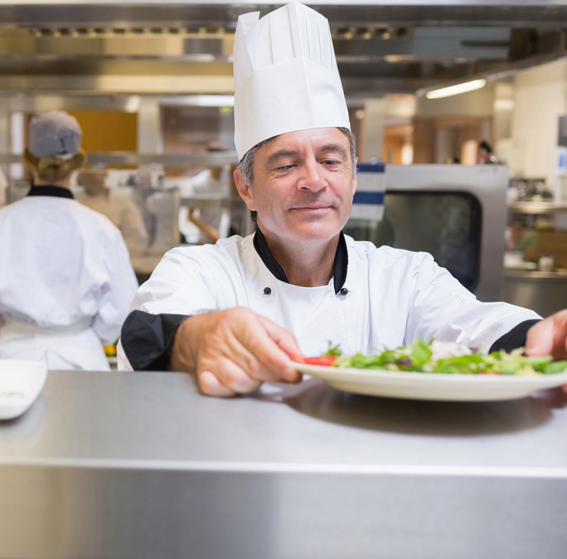 A chef in a white uniform and hat is carefully plating a salad.