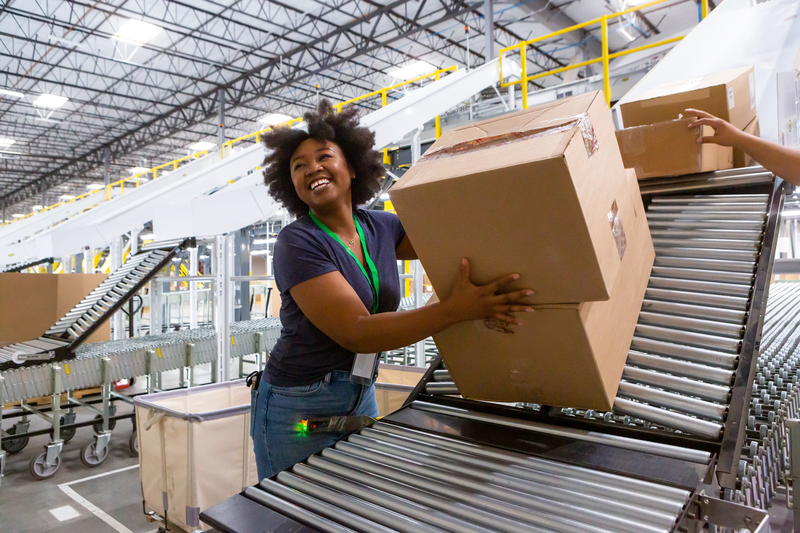 A smiling woman wearing a green lanyard works in a warehouse, lifting a box onto a conveyor belt.