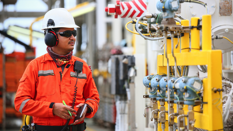An oil rig worker wearing a hard hat and safety glasses stands next to a large yellow pipe.